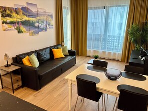 A modern living room with a black leather sofa, a small table, a dining table with chairs, and a large window with curtains.