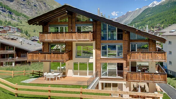 Un chalet en bois à plusieurs étages, doté de balcons, de grandes fenêtres et d’une terrasse aménagée avec du mobilier d’extérieur.