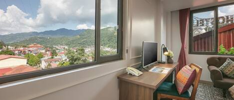 Habitación con un amplio ventanal que ofrece vistas a un pueblo y montañas. Hay un escritorio con computadora, una silla y una mesita con lámpara.