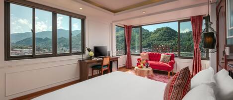 A hotel room with a large bed, a desk with a computer, a red sofa, and large windows offering a mountain view.