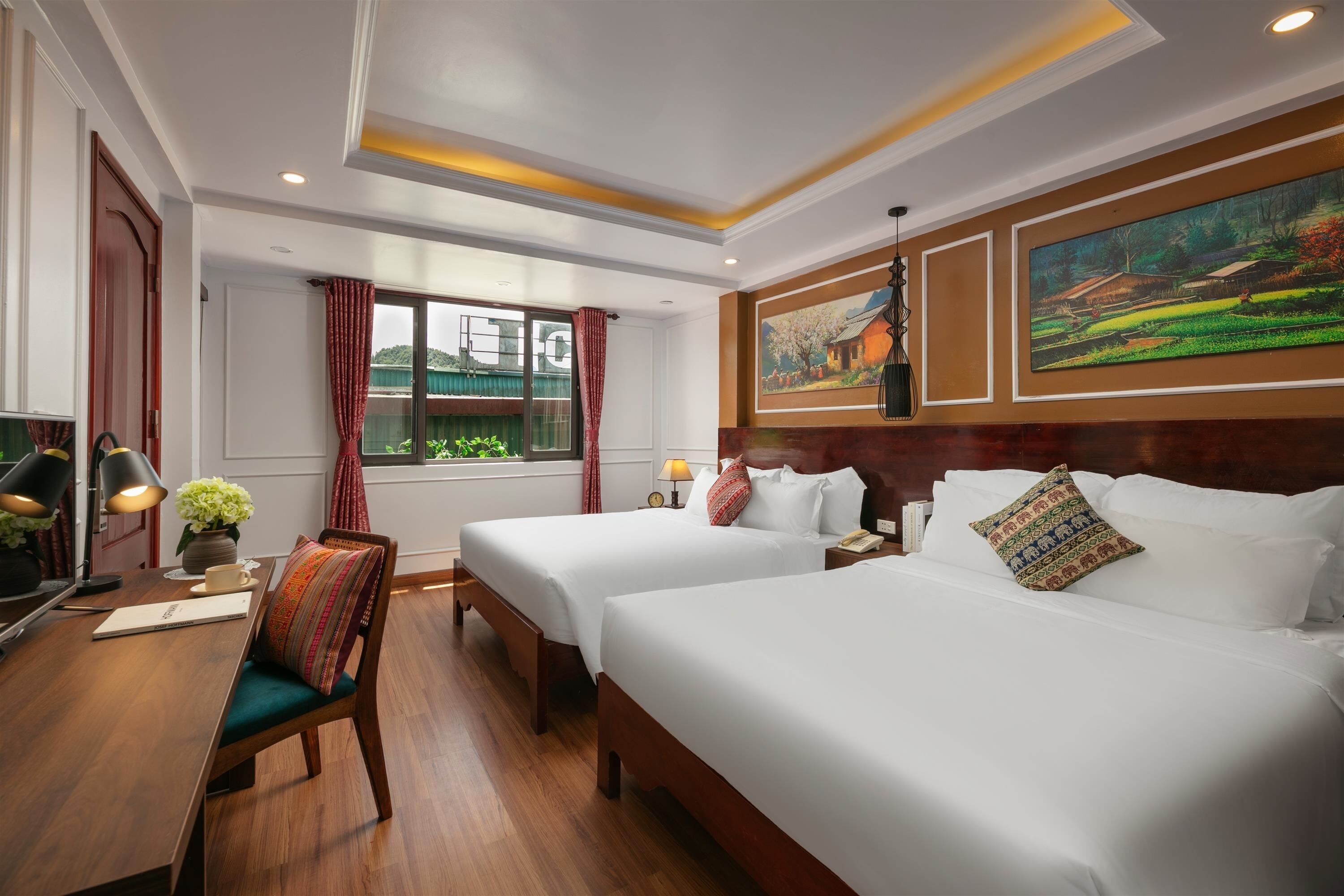 A hotel room with two beds, a desk with a lamp, a chair, a window with curtains, and paintings on the walls.