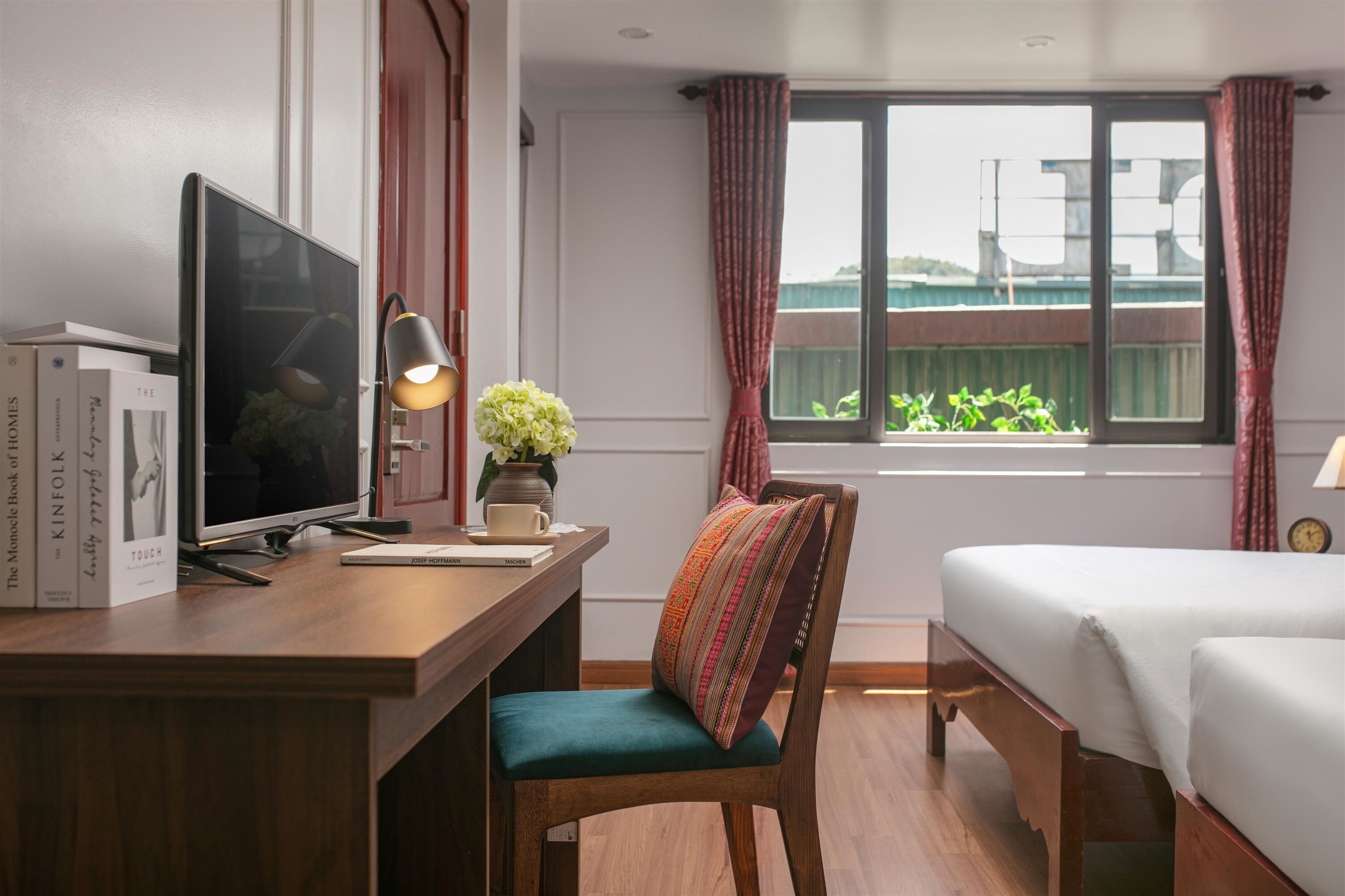 A hotel room with a desk, a chair, a bed, a window with curtains, and a view of greenery outside.