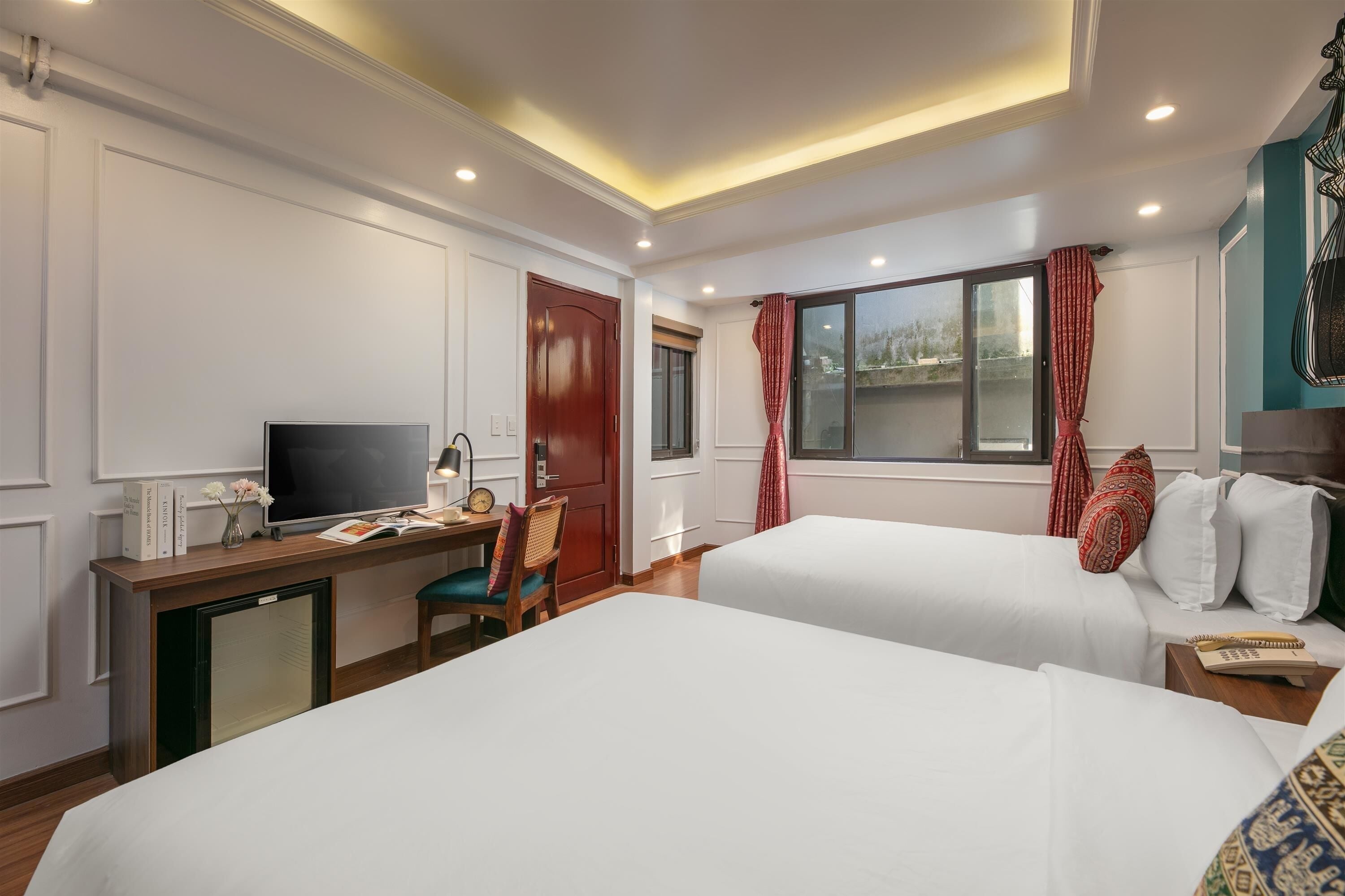 A hotel room with two beds, a desk with a TV, a small fridge, and a window with red curtains.