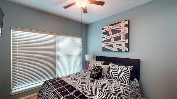 A bedroom with a bed, a ceiling fan, a window with blinds, a wall-mounted abstract painting, and a bedside lamp.