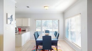 A dining area with a table and chairs, a kitchen in the background, and large windows with blinds.