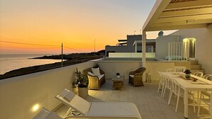 A rooftop terrace with lounge chairs, a dining table, and a view of the sea at sunset.