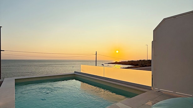 A swimming pool with a view of the sunset over the sea, a tiled deck, and a green cushion.