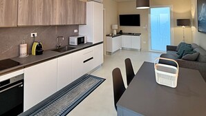 A modern kitchen with white cabinets, a microwave, and a coffee maker. There is a dining area with a table and chairs, and a sofa in the background.