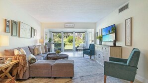 Amplia sala de estar con un conjunto de sofás, una mesa de centro y un televisor. Se ve un jardín a través de una puerta de cristal.