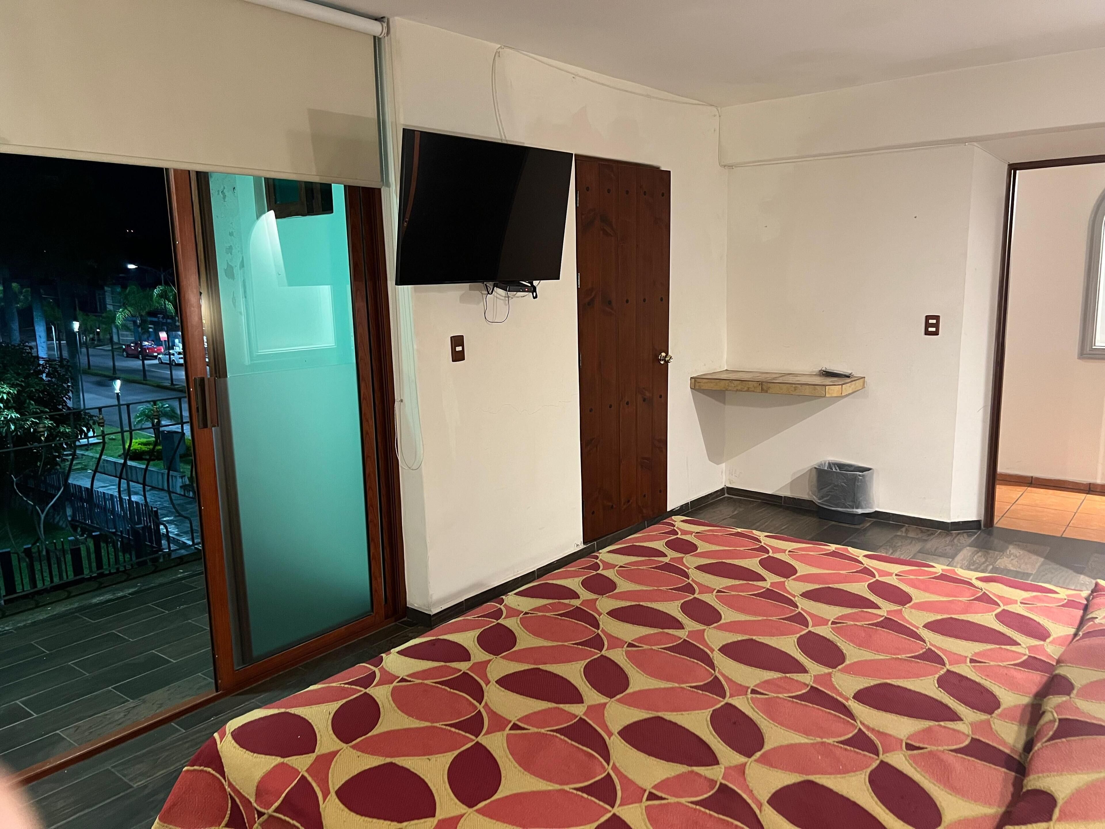 A room with a sliding glass door, a flat-screen TV mounted on the wall, a wooden door, a small shelf, and a patterned rug.