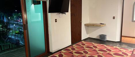 A room with a sliding glass door, a flat-screen TV mounted on the wall, a wooden door, a small shelf, and a patterned rug.