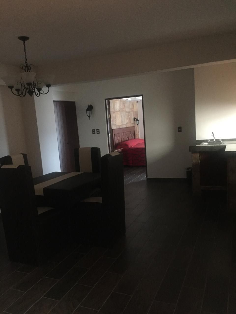 Ein Wohnzimmer mit Sofagarnitur, einer Chandelieber und Blick in ein Schlafzimmer mit rotem Bettbezug.