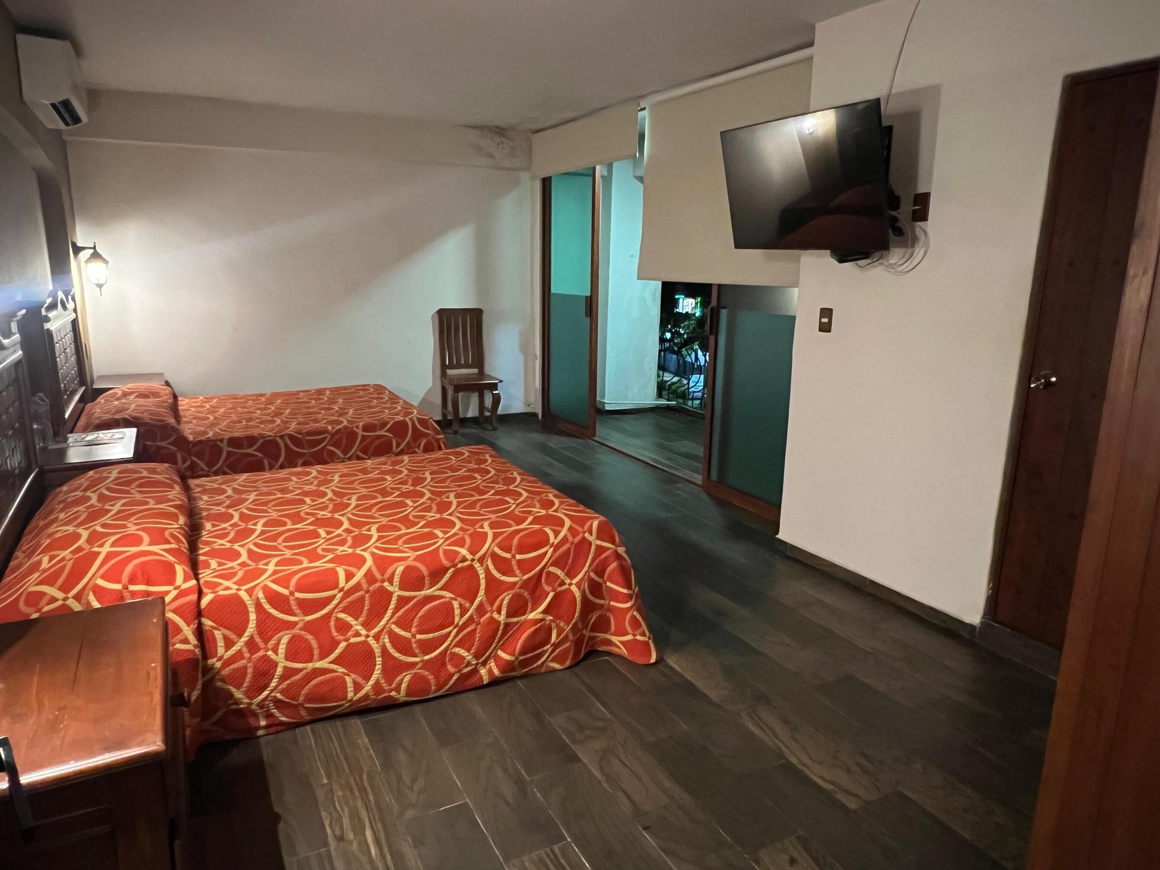 Habitación de hotel con dos camas, un televisor montado en la pared, una silla y una puerta que da a un balcón.