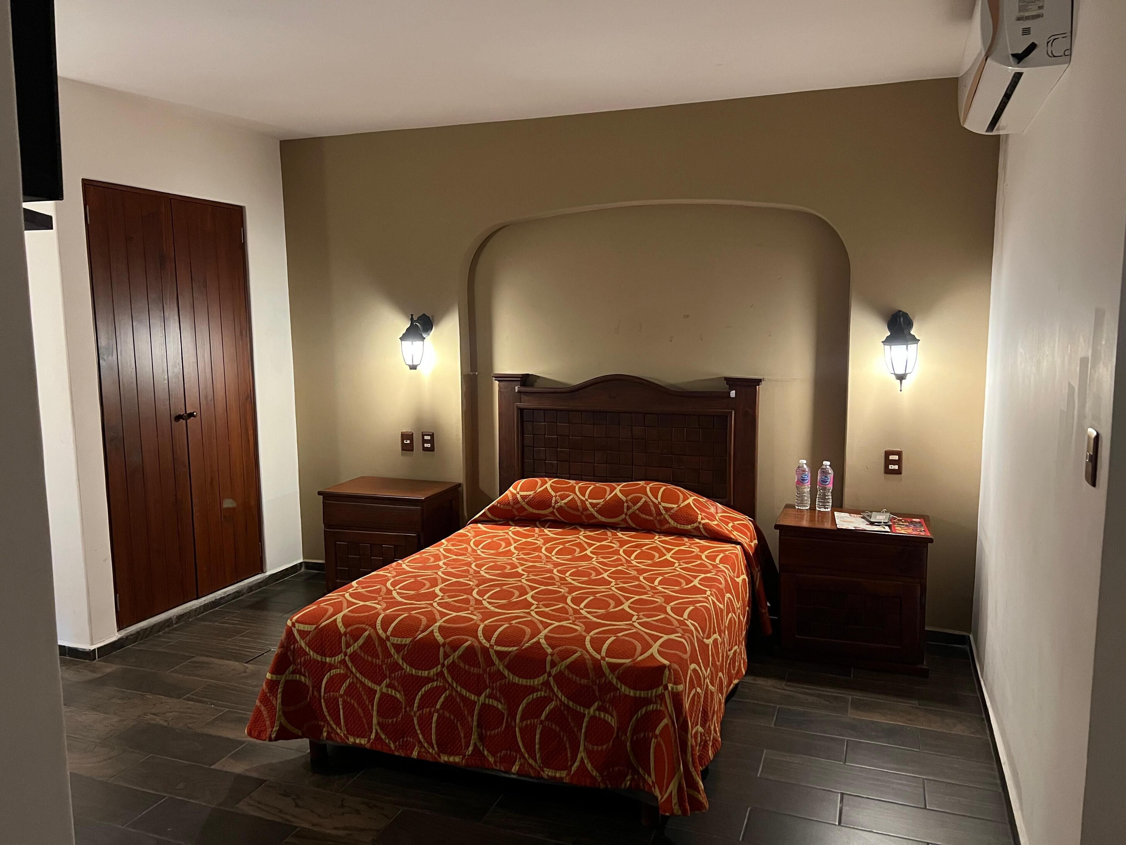 A hotel room with a bed, two bedside tables, a nightstand, a wooden wardrobe, and wall-mounted lamps.