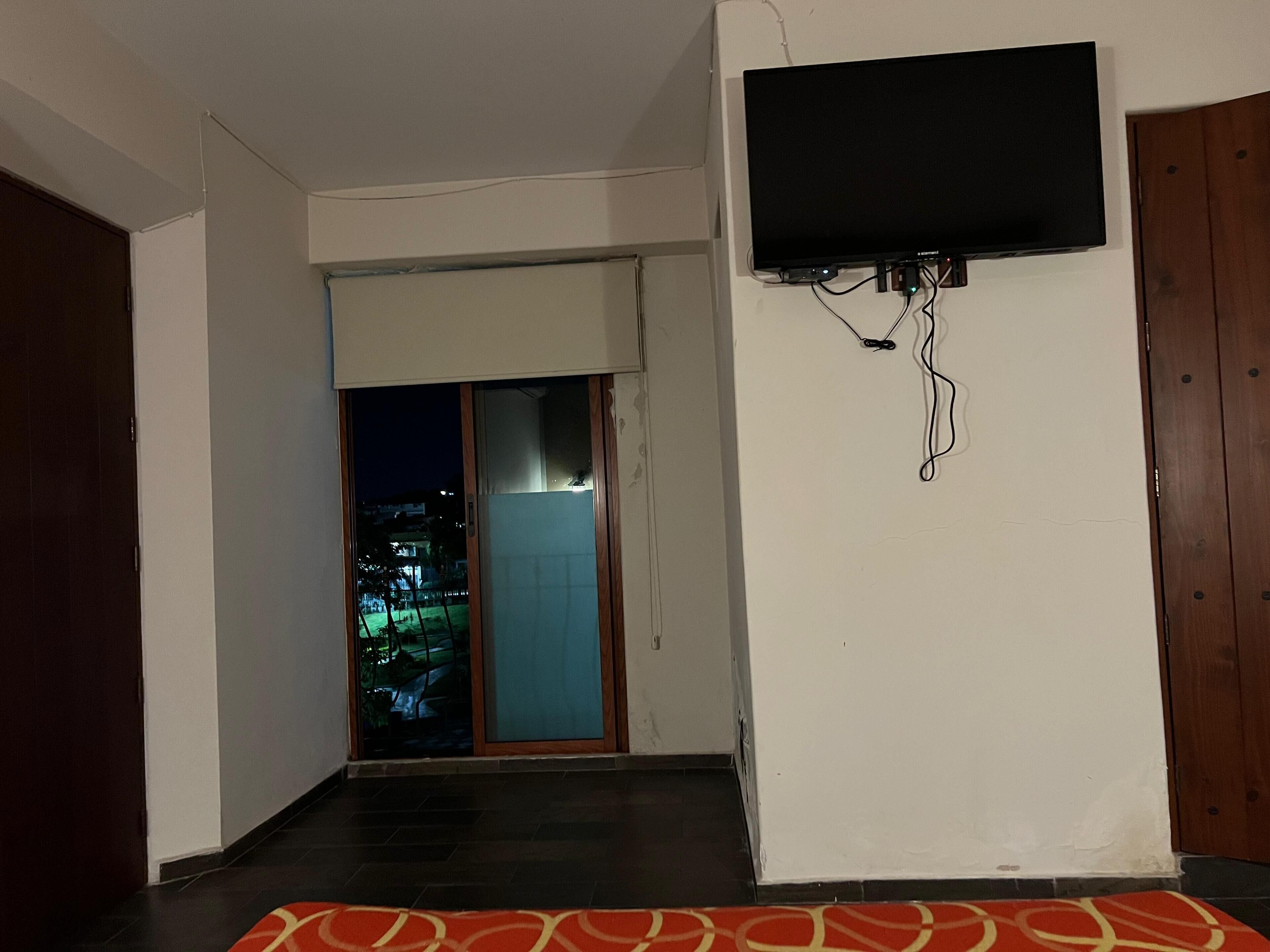 A room with a TV mounted on the wall, a sliding glass door leading to a balcony, and a red patterned rug on the floor.