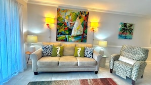 Una sala de estar con un sofá beige, un sillón estampado y dos mesitas de noche con lámparas. Un cuadro abstracto colorido y un cuadro con una palmera decoran las paredes.