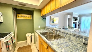 Una cocina con paredes verdes, armarios de madera y salpicadero de azulejos. Se observa un lavamanos doble, una estufa y un cubo de basura.