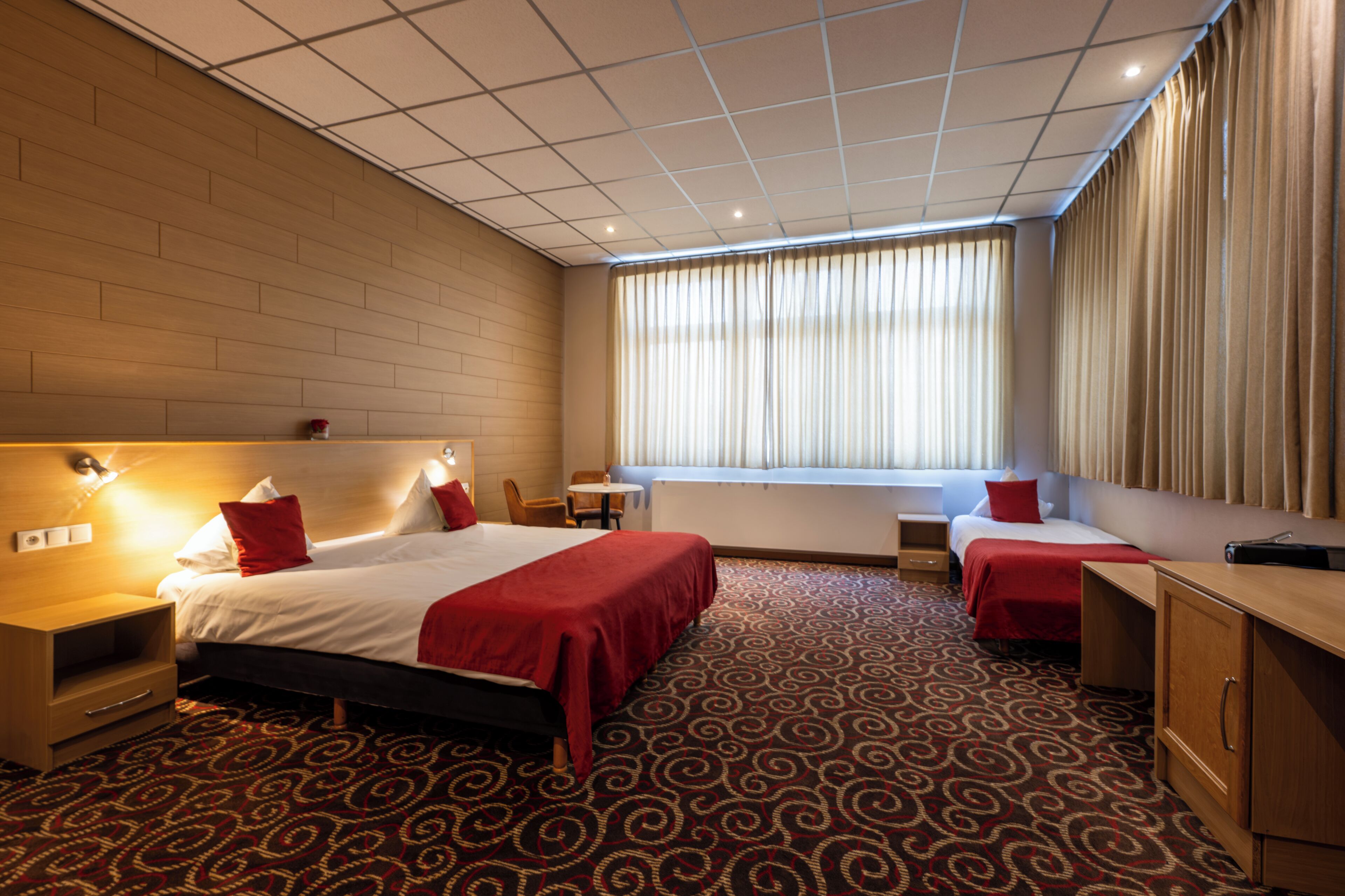 A hotel room with a large bed, a smaller bed, a desk, a chair, a window with curtains, and a patterned carpet.