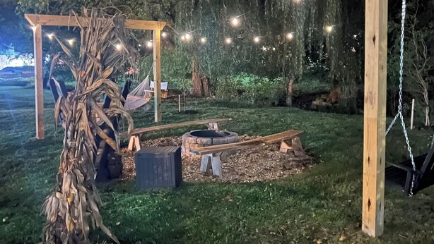 Eine rustikale Außenanlage mit einer Holzpergola, Lichterketten, einer Feuerstelle und einem Baum.