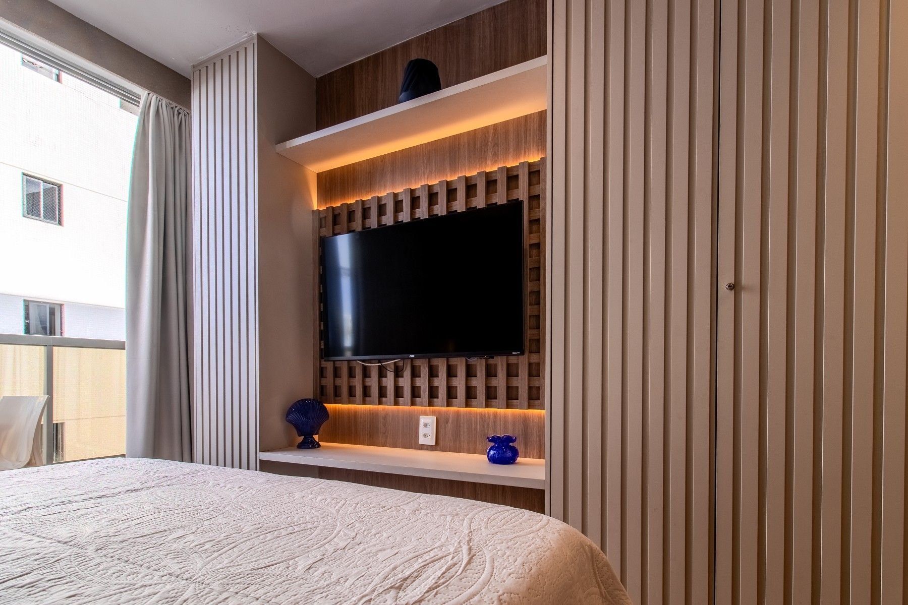 Habitación de hotel con una cama grande, un televisor montado y un estante empotrado con adornos.
