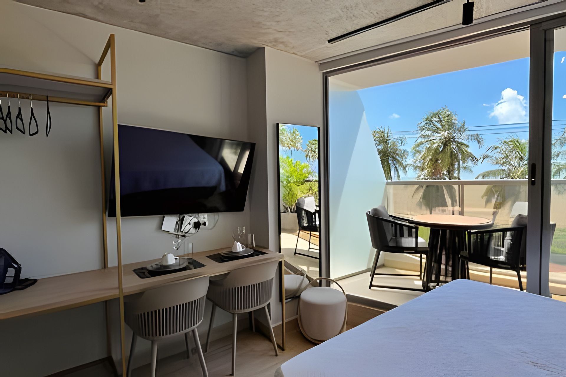Una habitación de hotel con cama, escritorio con dos sillas, televisor montado en la pared y balcón con mesa y sillas.