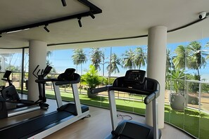 A gym with treadmills and exercise equipment, large windows, and a view of palm trees. - Setai Yach (Joao Pessoa)