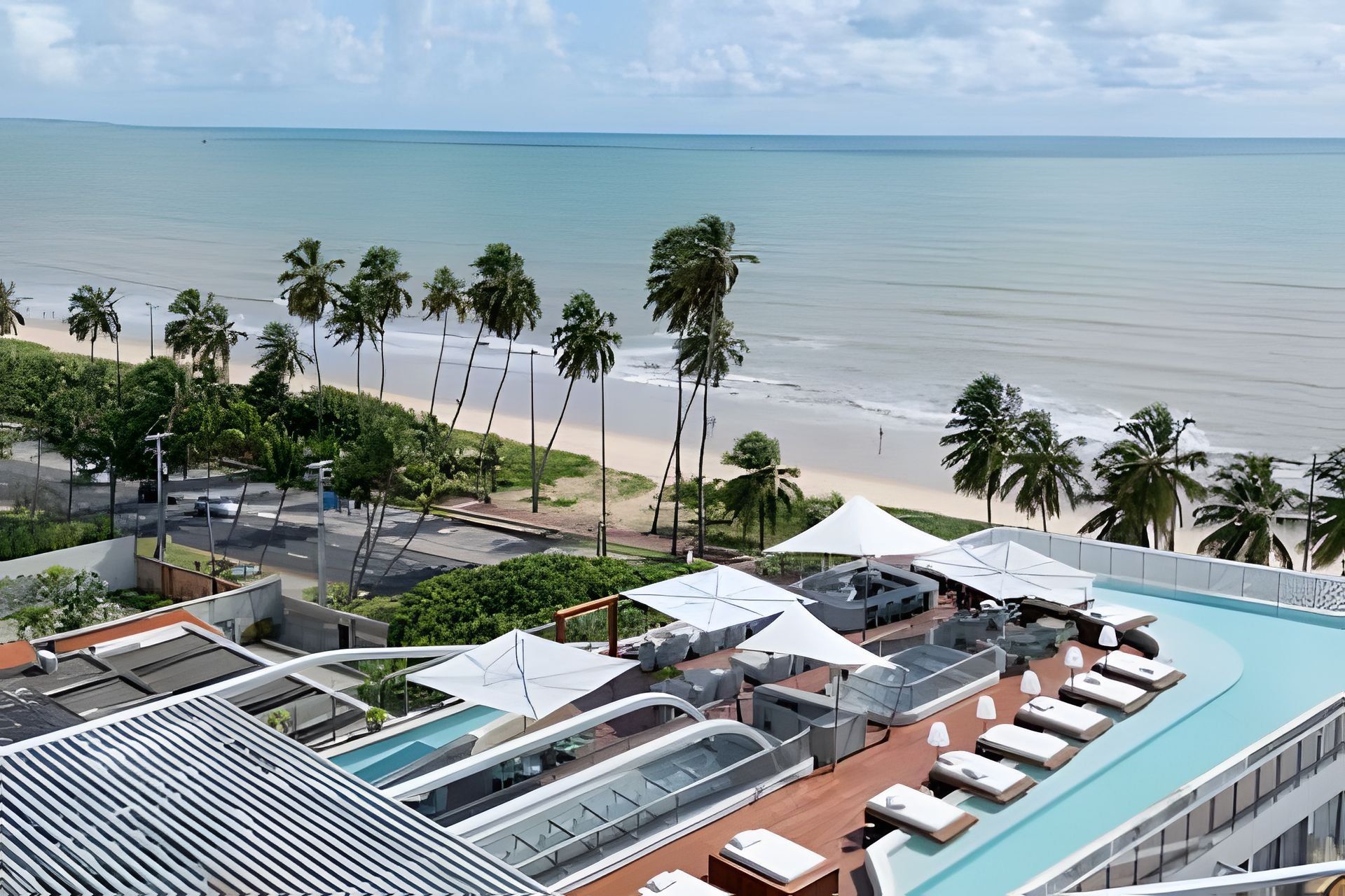 Zona de piscina en la azotea con sillones y sombrillas, con vistas a una playa con palmeras y cielo despejado.