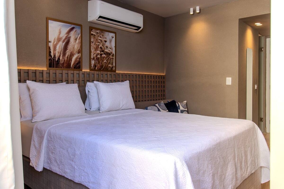 A hotel room with a bed, white bedding, an air conditioning unit, and two wall-mounted pictures.