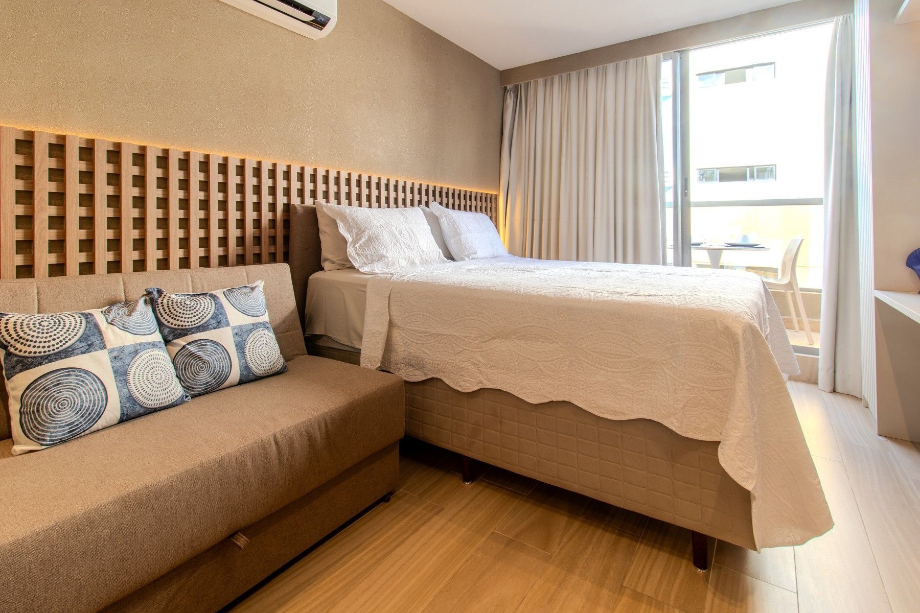 Habitación de hotel con cama, sofá, cabecero de madera y un ventanal con cortinas.