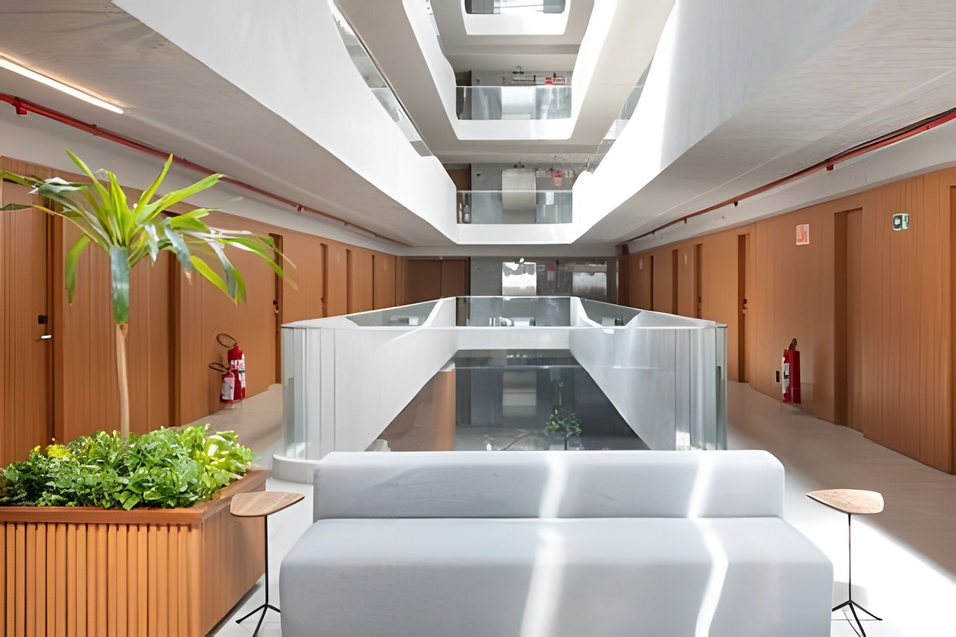 Un moderno vestíbulo de hotel con un atrio de cristal central, paneles de madera en las paredes, una zona de estar con sofá y una pequeña maceta de madera con plantas.