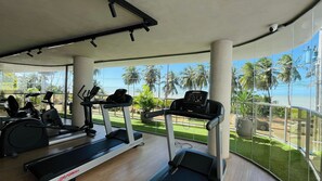 Ein Fitnessstudio mit Laufbändern, Ergometer und Rudergerät, mit großen Fenstern und Blick auf Palmen und einen klaren Himmel.