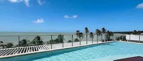 A rooftop pool with clear blue water, a wooden deck, and a view of the ocean.