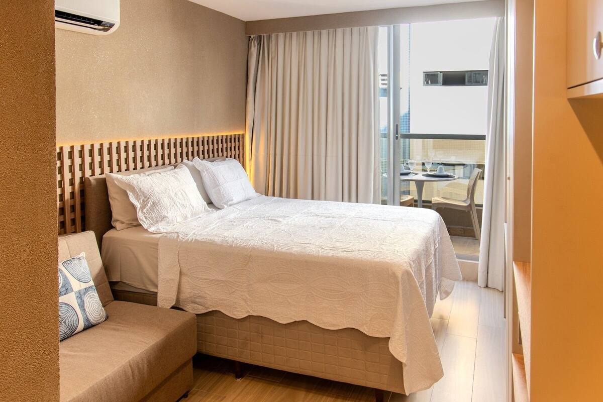 A hotel room with a bed, a sofa, a balcony with a table and chairs, and an air conditioning unit.