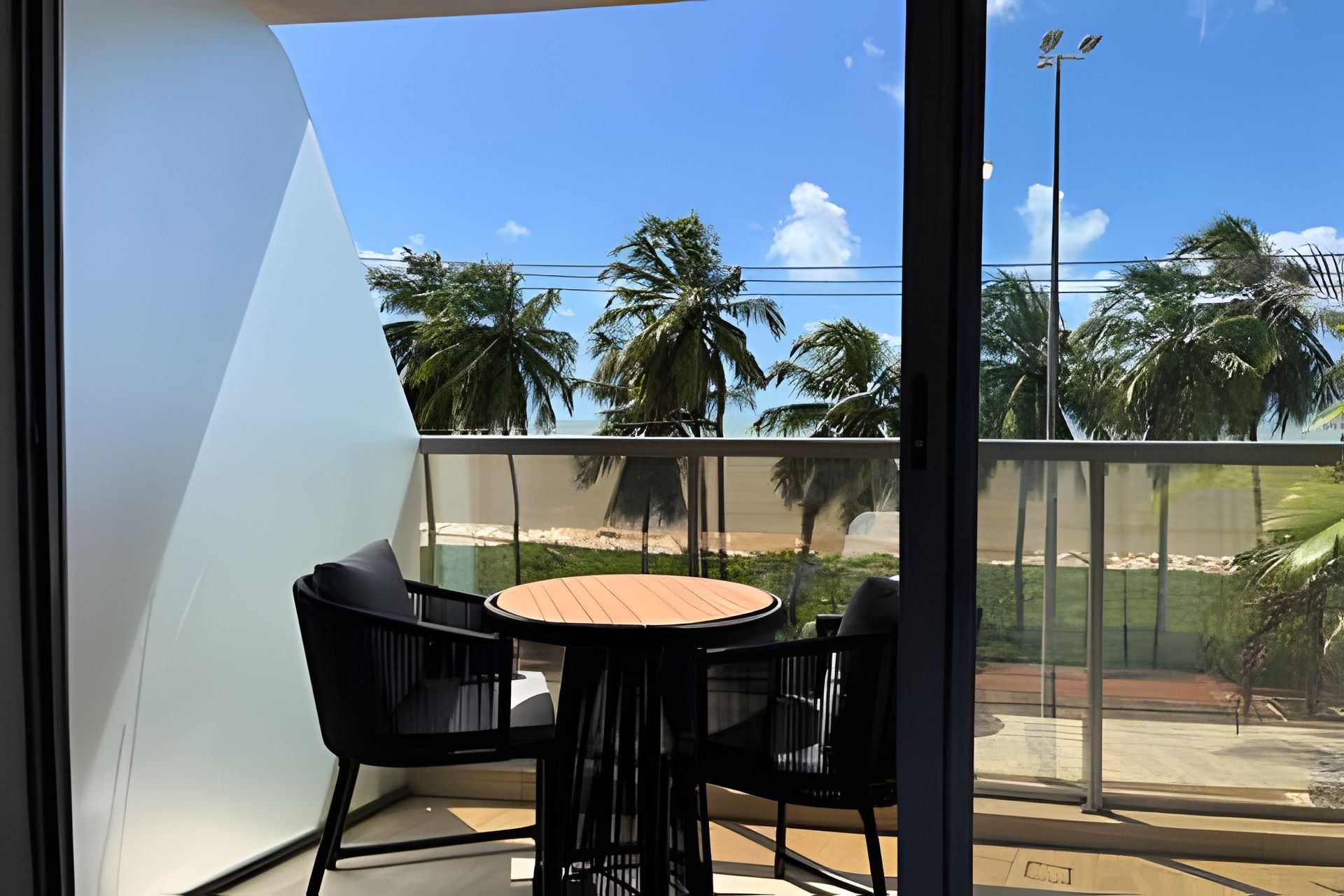 Varanda com mesa redonda e duas cadeiras, vista para uma praia com palmeiras e céu claro.