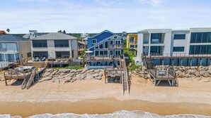 A beachfront property with multiple buildings, including a large blue house with a balcony and a smaller white house. There are wooden walkways leading to the beach and a sandy area with a rocky shoreline.