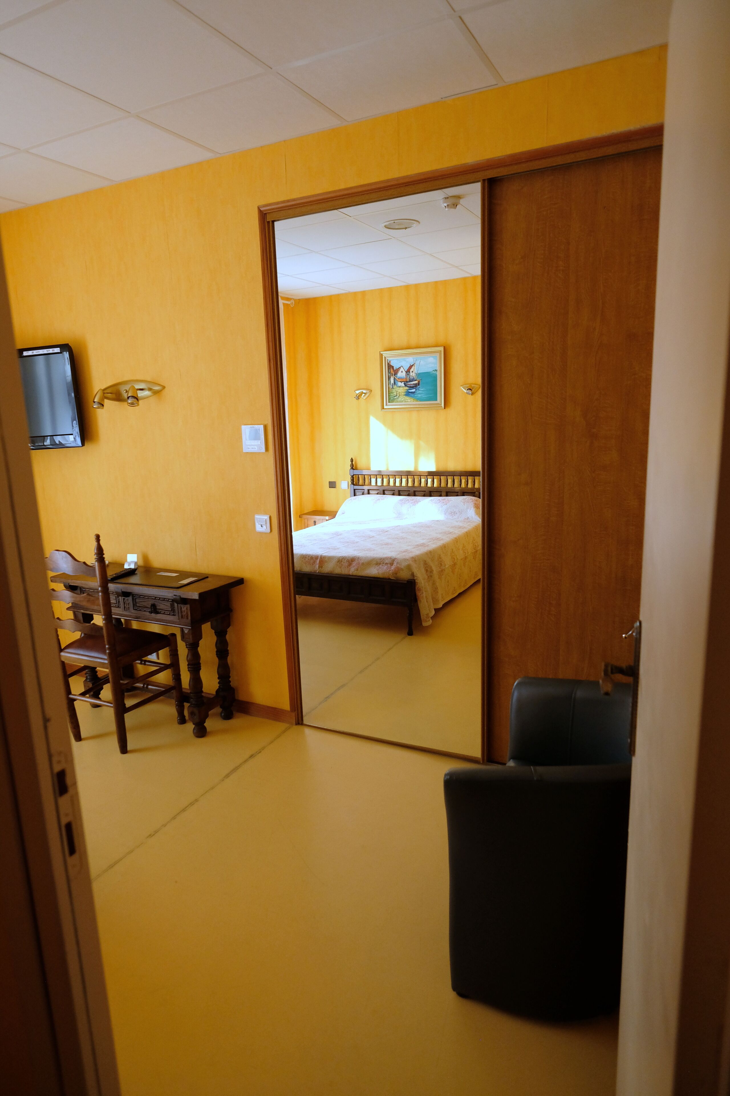 A hotel room with a wooden desk, a chair, a bed, and a mirror reflecting the room.
