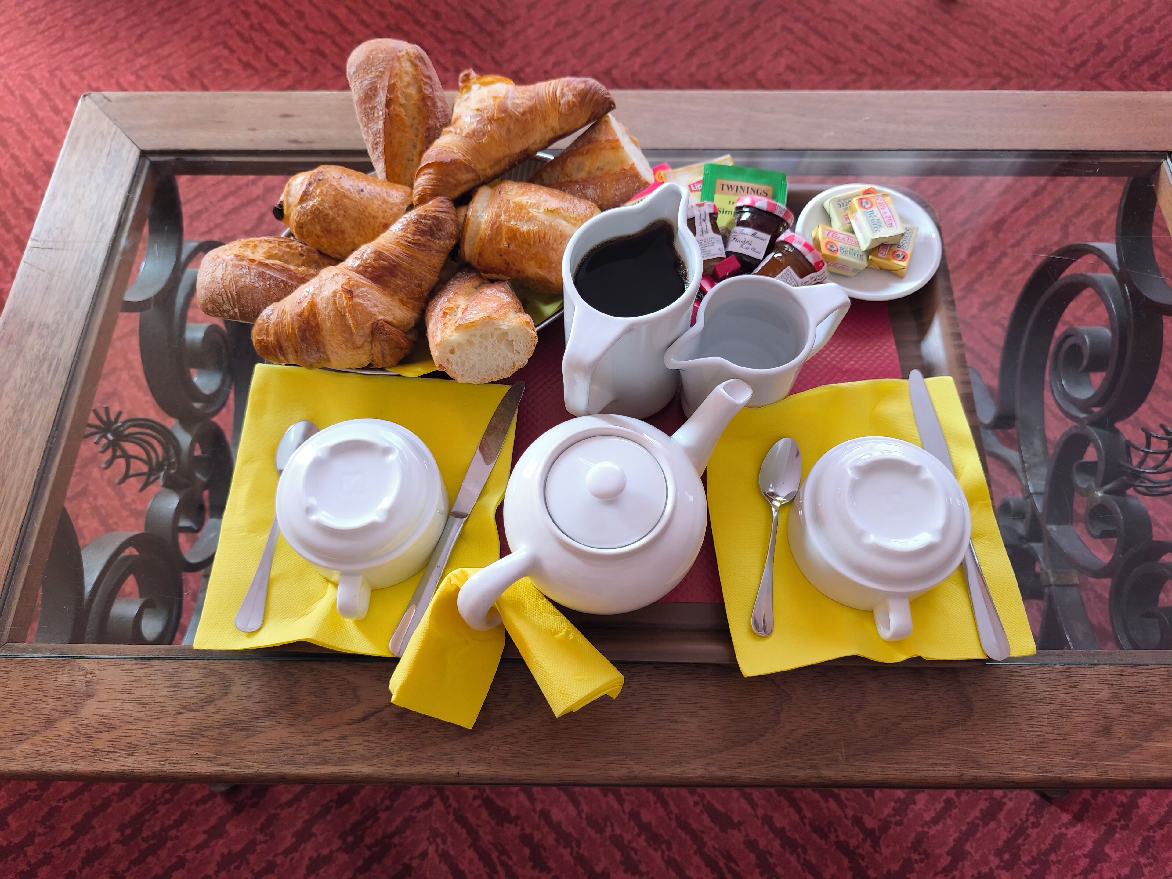 A breakfast tray with croissants, jam, butter, and coffee on a glass table.
