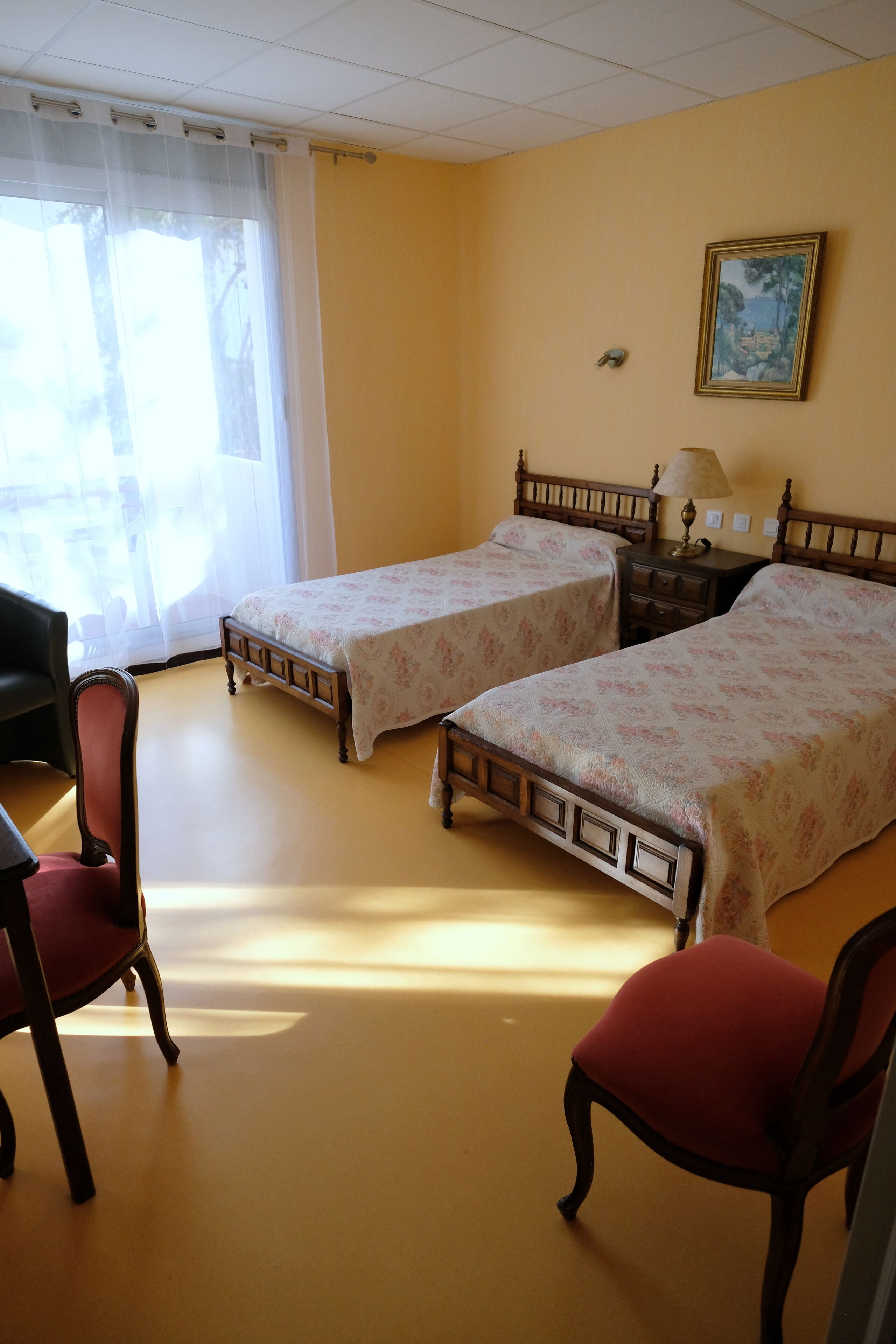 Een hotelkamer met twee enkele bedden, een bureau, een stoel en een raam met dunne gordijnen.