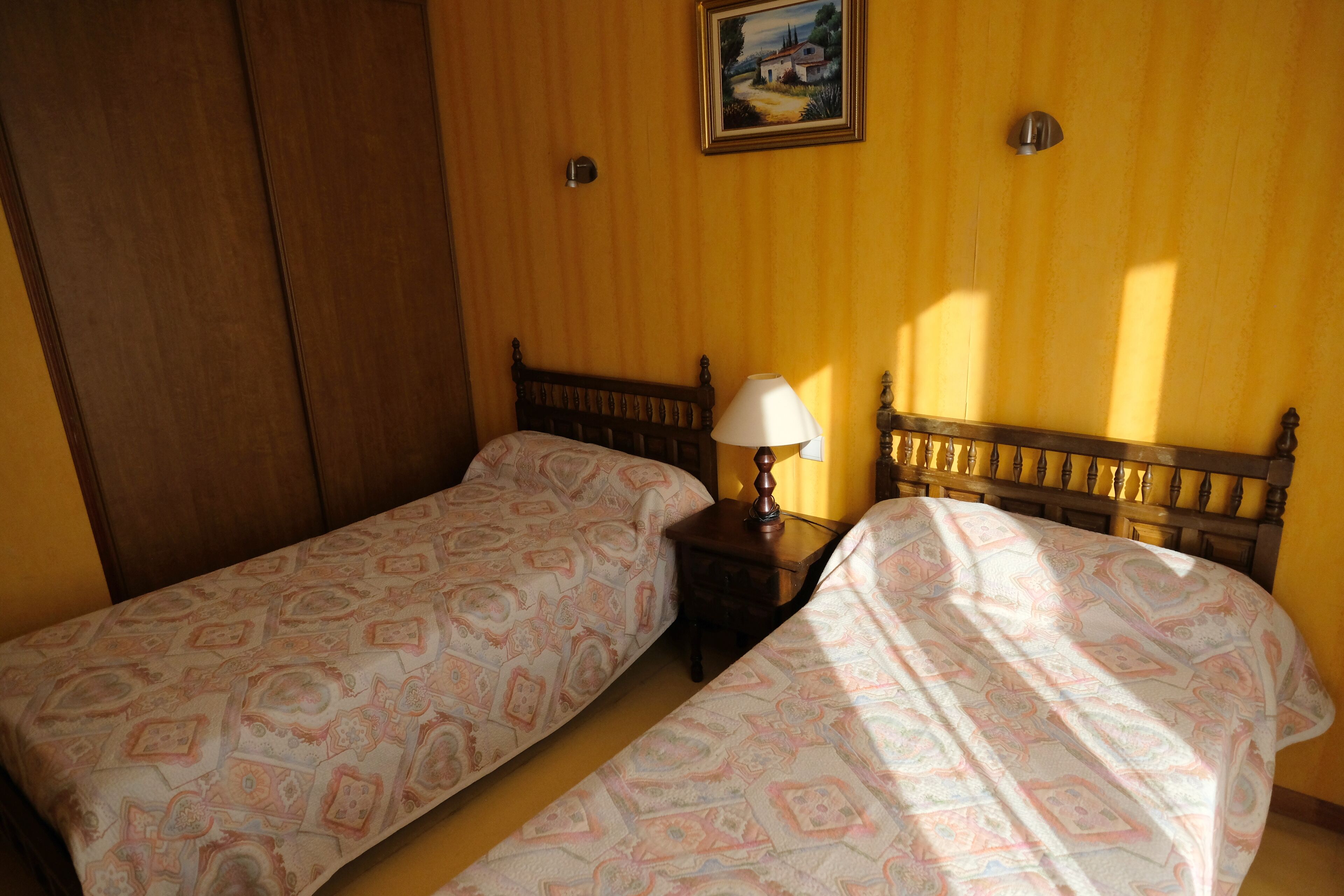 A room with two single beds, wooden headboards, a nightstand with a lamp, and a painting on the wall.