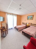 Quarto de hotel com duas camas de solteiro, uma escrivaninha com cadeira, uma poltrona vermelha, uma televisão e um quadro na parede.