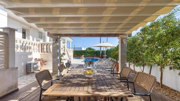 En uteplats med ett träbord, flätade stolar, en pool och ett takförsett område med en pergola.