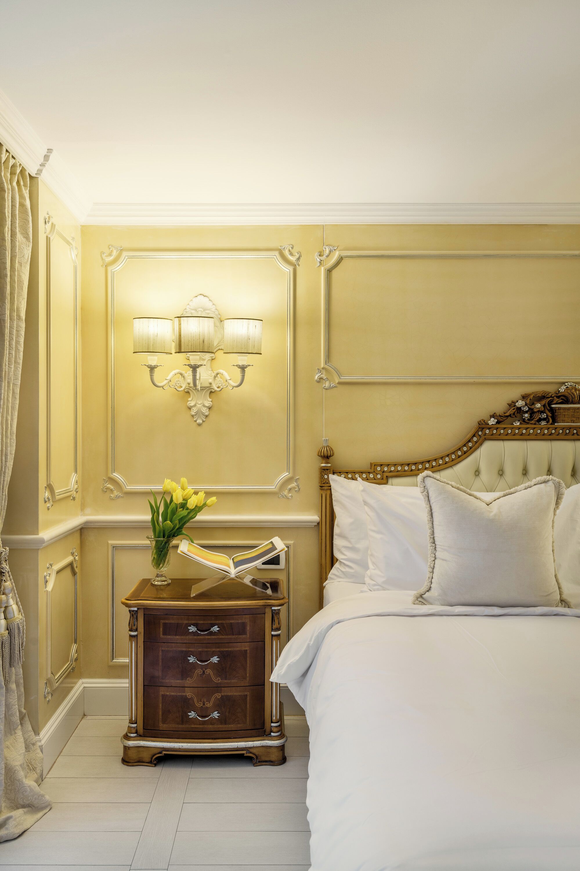 Un dormitorio con cama, mesita de noche, lámpara y un florero con flores.