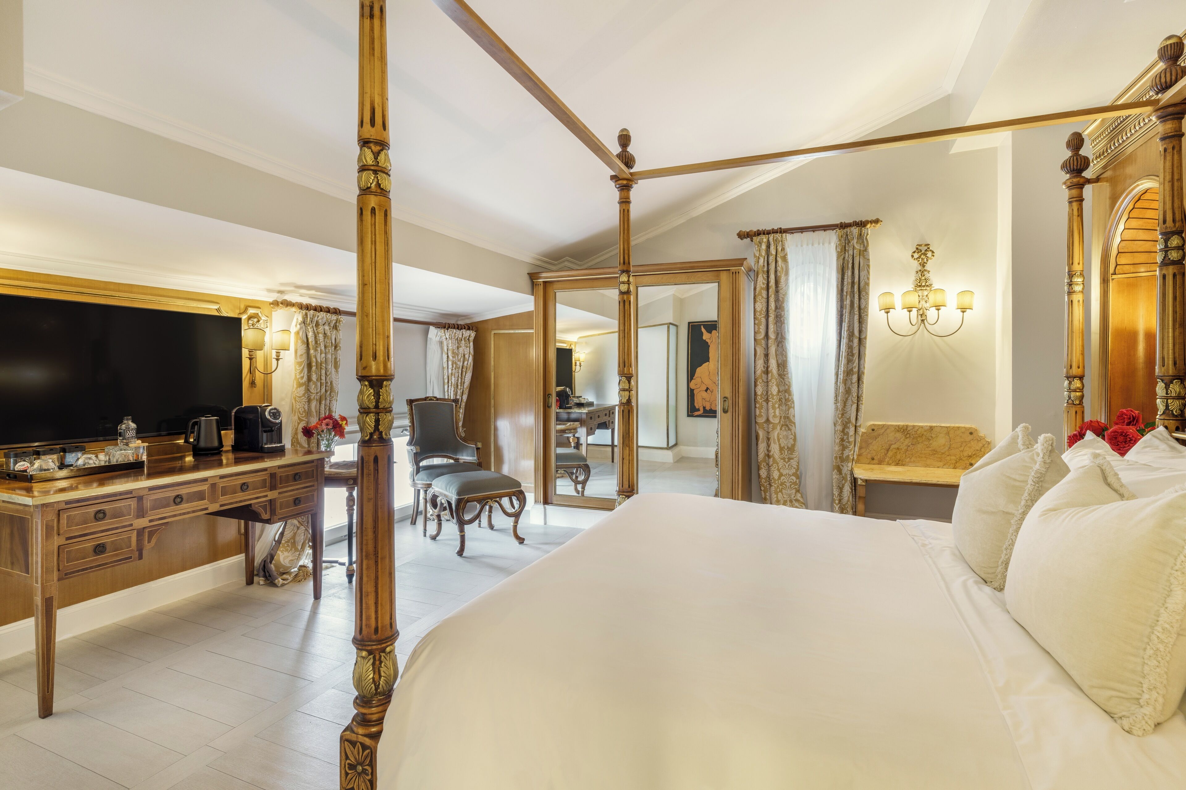 A luxurious bedroom with a four-poster bed, a large flat-screen TV, a wooden desk, and a dining area visible through a mirror.