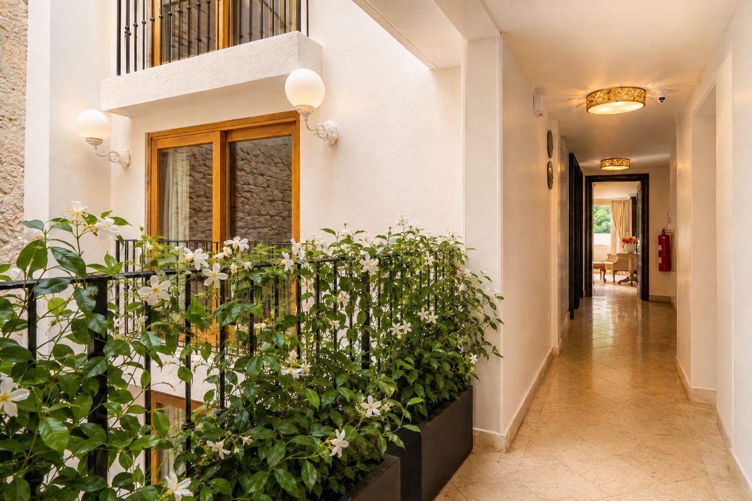 Un pasillo de hotel con una puerta de madera, una barandilla de metal negro decorada con flores blancas y un piso de baldosas.
