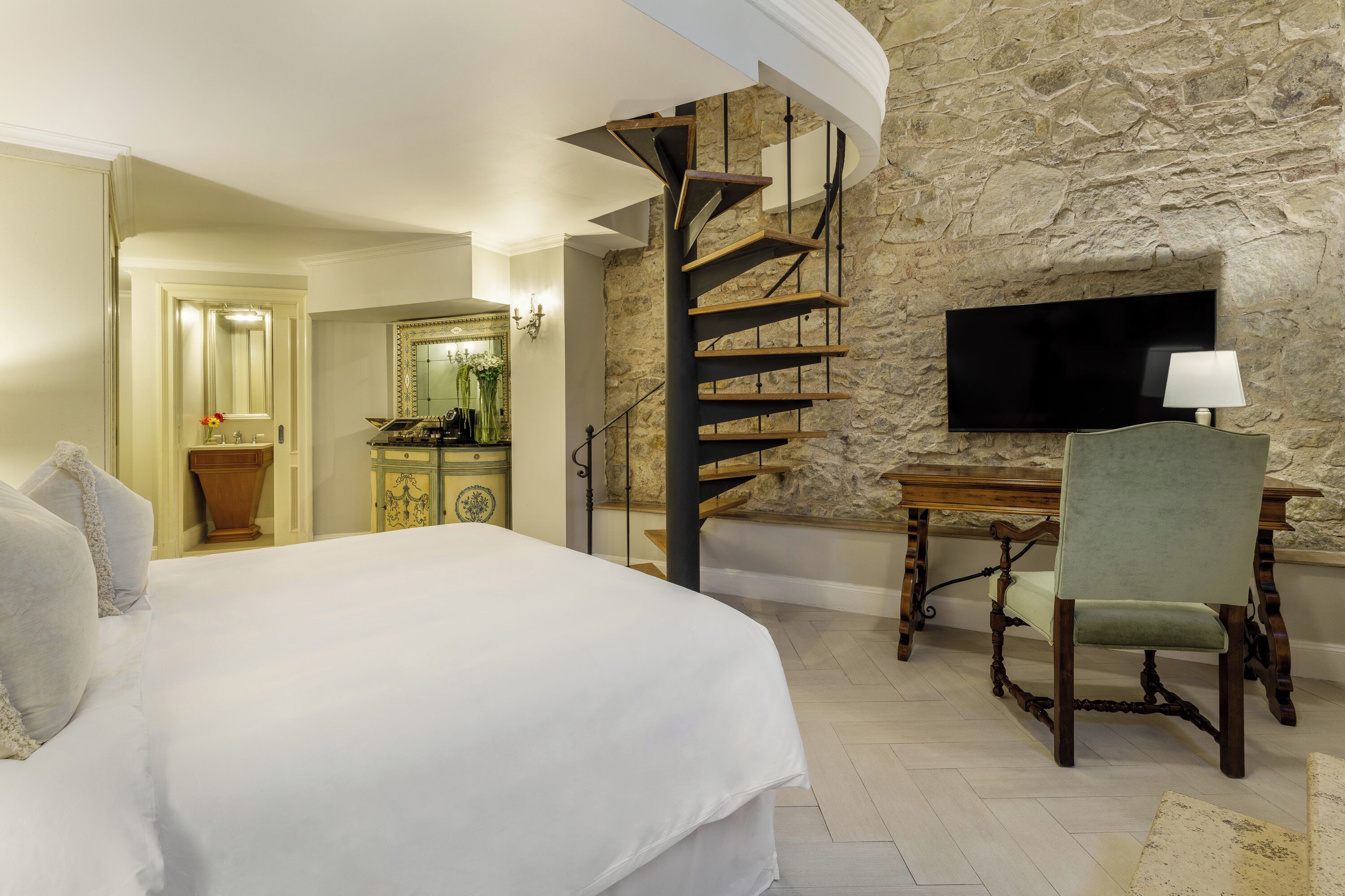 Un dormitorio con una cama grande, un escritorio con silla, una pared de piedra y un televisor montado en la pared.