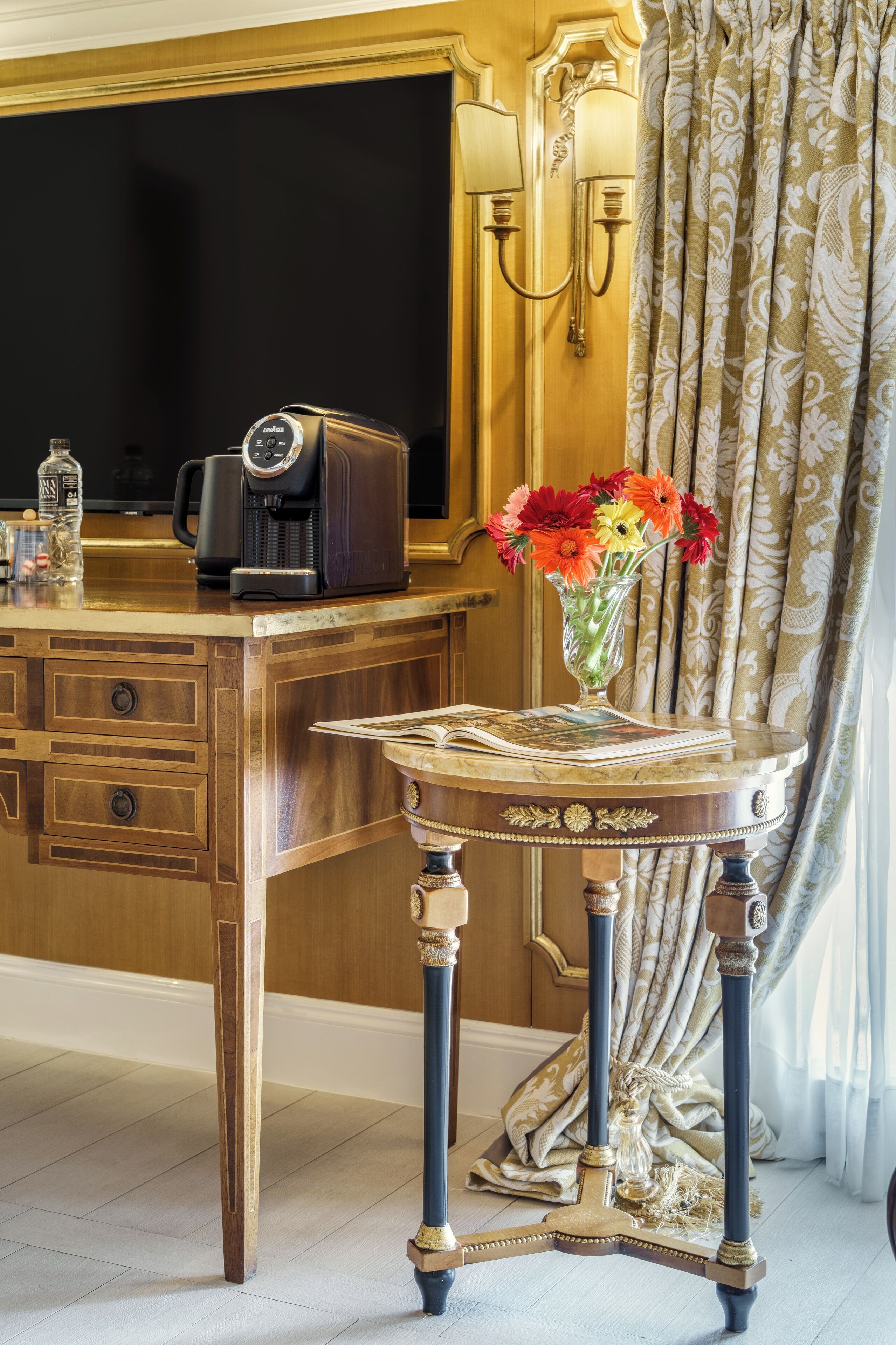 A wooden desk with a coffee maker, a vase with flowers, and a magazine.