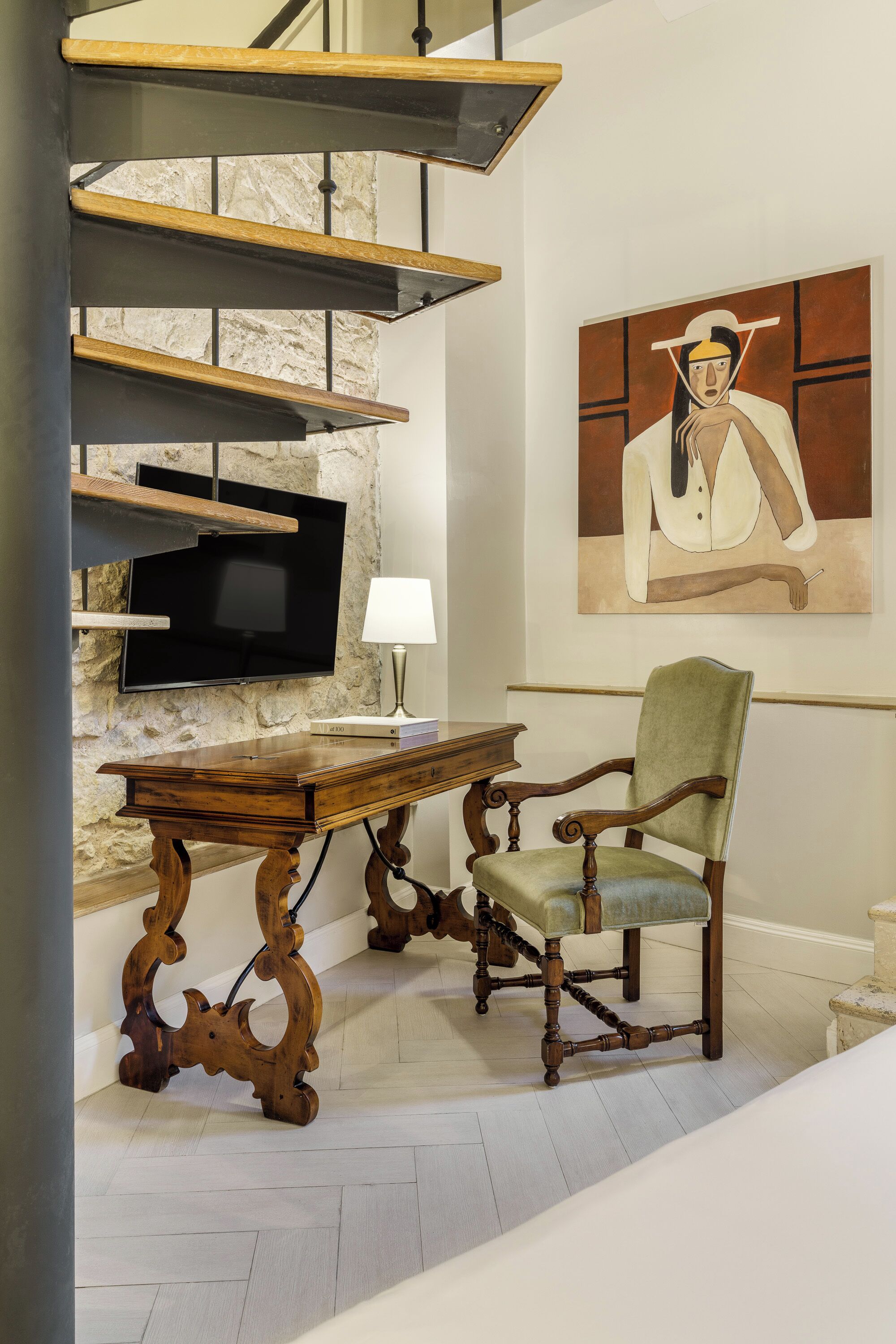 A staircase with wooden steps and metal railings, a flat-screen TV mounted on a stone wall, a wooden table with a lamp, and a green upholstered chair.