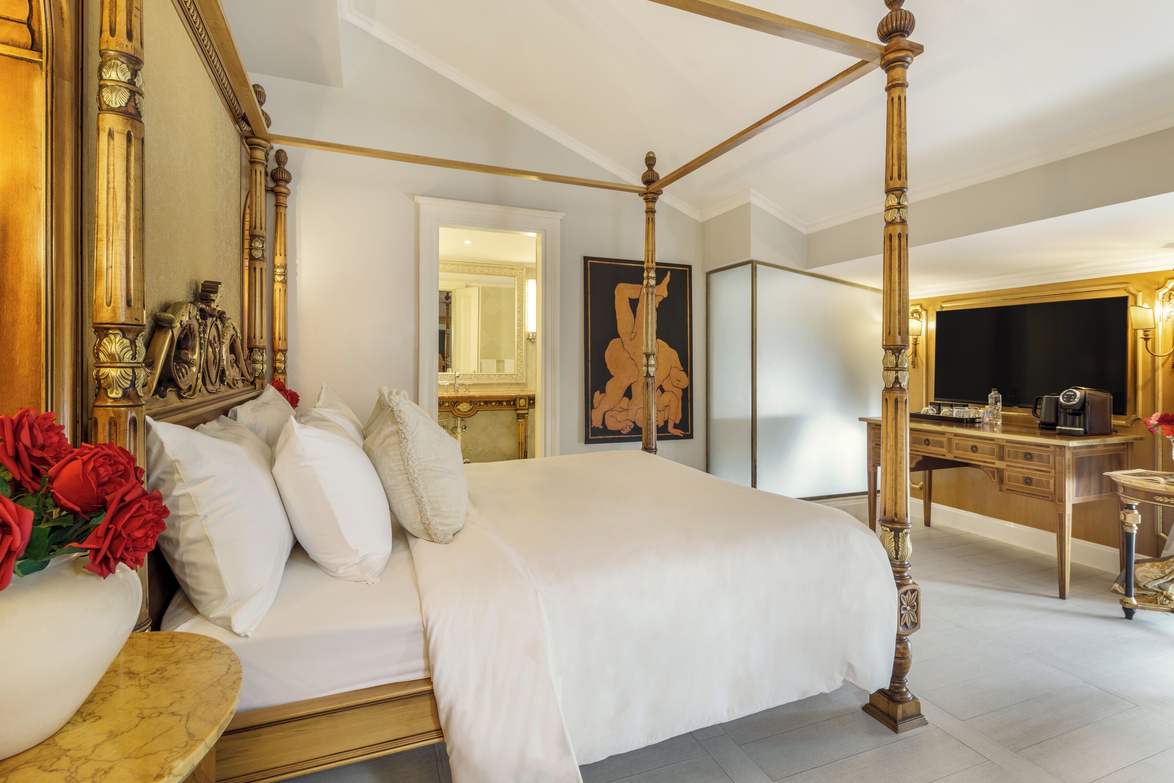 A four-poster bed with white linens, a marble bedside table with red roses, a wooden desk with a mirror, and a framed artwork on the wall.