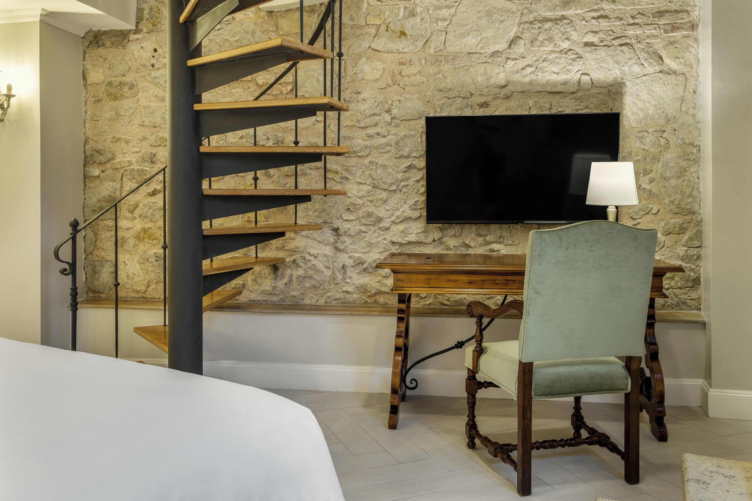 A bedroom with a stone wall, a wooden staircase, a flat-screen TV, a wooden desk, and a chair.
