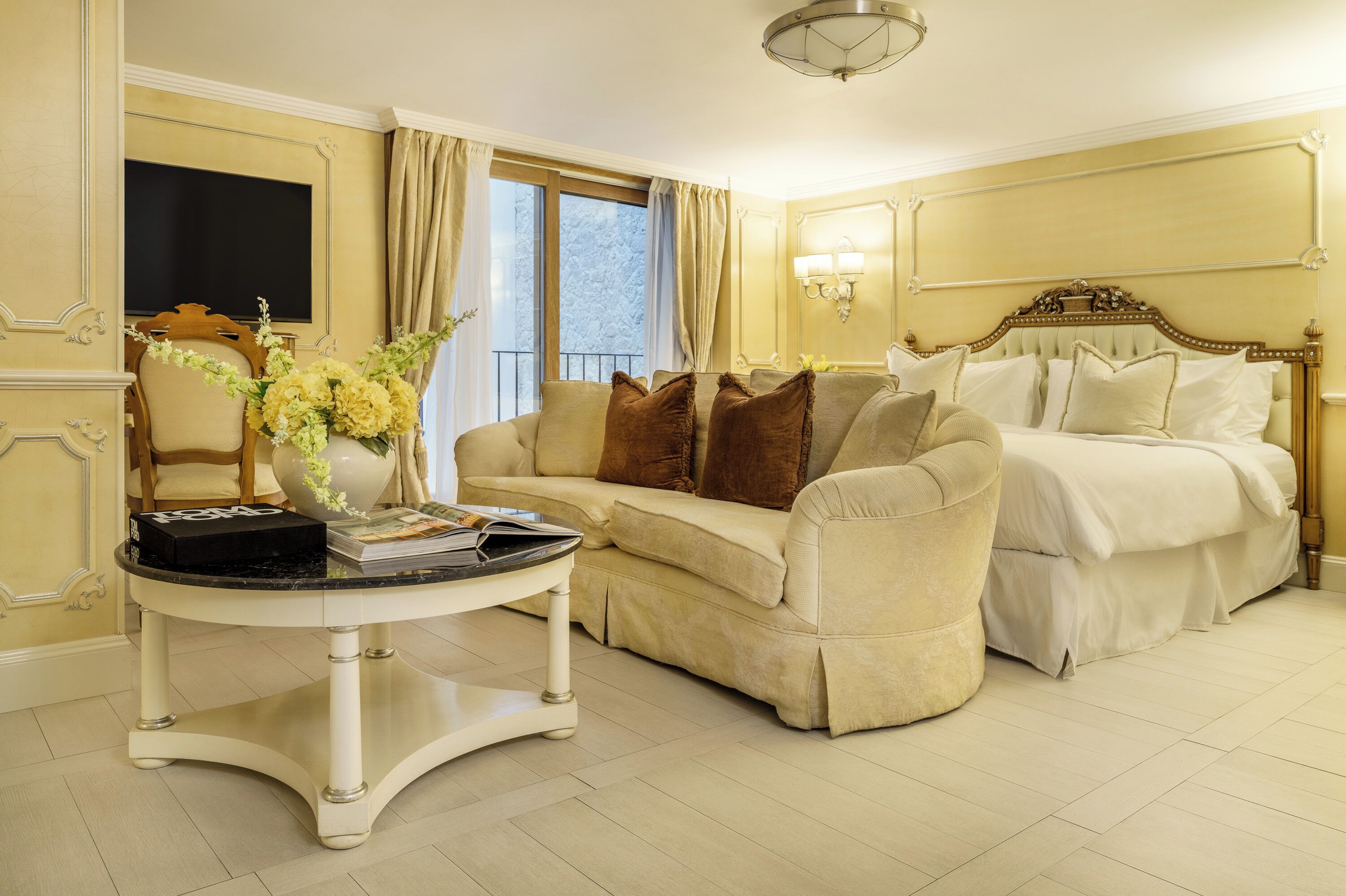 Ein Hotelzimmer mit beiger Couch, einem weißen Bett, einem runden Tisch mit Blumenvase und einem großen Fenster mit Vorhängen.